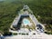 Aerial view of a large resort complex surrounded by lush greenery. The centerpiece is a large swimming pool lined with lounge chairs, nestled within well-maintained gardens. The structures are built in a U-shape, creating an inviting central area. Adjacent to the resort is a pristine white sandy beach with additional sun loungers and shaded areas, indicating a tropical setting.
