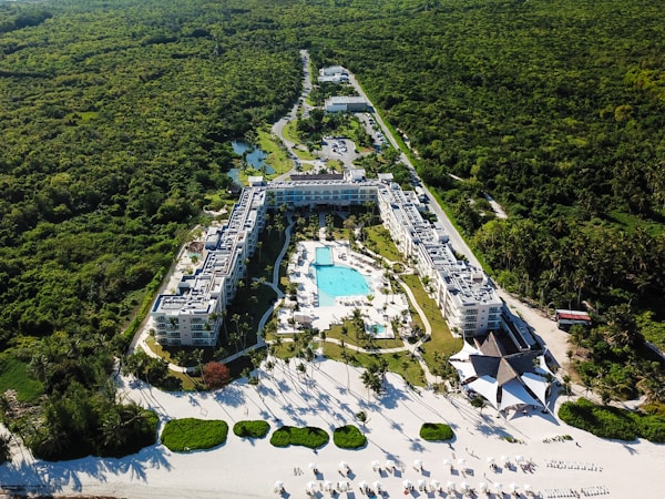 Aerial view of a large resort complex surrounded by lush greenery. The centerpiece is a large swimming pool lined with lounge chairs, nestled within well-maintained gardens. The structures are built in a U-shape, creating an inviting central area. Adjacent to the resort is a pristine white sandy beach with additional sun loungers and shaded areas, indicating a tropical setting.
