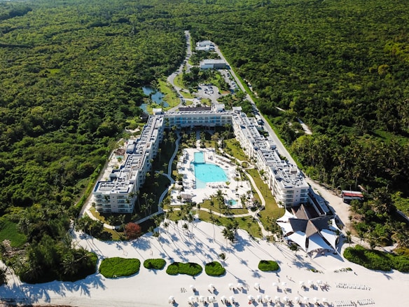 Aerial view of a large resort complex surrounded by lush greenery. The centerpiece is a large swimming pool lined with lounge chairs, nestled within well-maintained gardens. The structures are built in a U-shape, creating an inviting central area. Adjacent to the resort is a pristine white sandy beach with additional sun loungers and shaded areas, indicating a tropical setting.