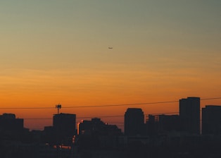 A city skyline with tall buildings silhouetted against a vibrant dusk or dawn sky. An airplane is seen flying across the sky, while power lines stretch across the scene.