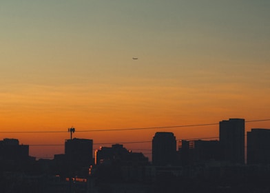 A city skyline with tall buildings silhouetted against a vibrant dusk or dawn sky. An airplane is seen flying across the sky, while power lines stretch across the scene.