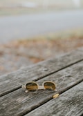 A pair of round sunglasses with clear frames rests on a weathered wooden picnic table. The background is blurred, showing hints of grass and a pathway.