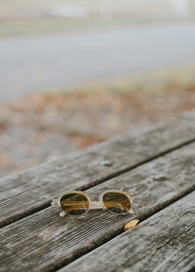 A pair of round sunglasses with clear frames rests on a weathered wooden picnic table. The background is blurred, showing hints of grass and a pathway.