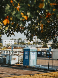 A portable toilet is situated in a parking area, surrounded by a chain-link fence. There are parking signs in the vicinity and several construction-related items are visible. The foreground has leafy branches partially obscuring the view, with residential buildings in the background.