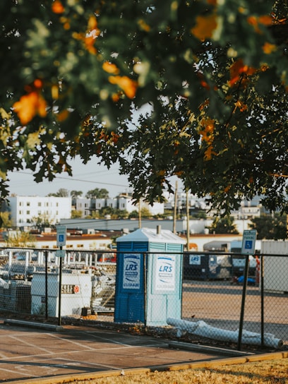 A portable toilet is situated in a parking area, surrounded by a chain-link fence. There are parking signs in the vicinity and several construction-related items are visible. The foreground has leafy branches partially obscuring the view, with residential buildings in the background.