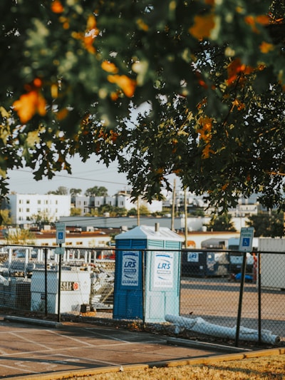 A portable toilet is situated in a parking area, surrounded by a chain-link fence. There are parking signs in the vicinity and several construction-related items are visible. The foreground has leafy branches partially obscuring the view, with residential buildings in the background.