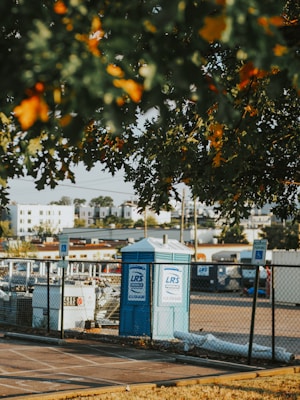 A portable toilet is situated in a parking area, surrounded by a chain-link fence. There are parking signs in the vicinity and several construction-related items are visible. The foreground has leafy branches partially obscuring the view, with residential buildings in the background.