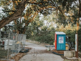 A portable toilet is positioned beside a construction site, surrounded by a chain-link fence. A small construction vehicle is visible next to the toilet, partially obscured by the surrounding foliage. The scene is framed by large, overhanging tree branches with green leaves.