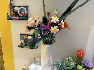 A collection of LEGO models featuring intricate designs including floral arrangements and miniature architectural landmarks. The flowers are crafted in various colors with detailed petals and leaves, displayed in a white vase. In the background, miniature LEGO buildings with signs reading 'London' and 'Paris' are mounted on a wall.