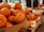 A variety of pumpkins are displayed in bulk containers and on tables. The pumpkins are in different sizes and colors, predominantly orange, with some white and multi-colored ones. The setting appears to be a marketplace or a grocery store, as suggested by the arrangement and the presence of baskets and a price sign.
