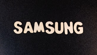 The white block letters spelling 'SAMSUNG' are arranged on a textured black surface, creating a high contrast visual.