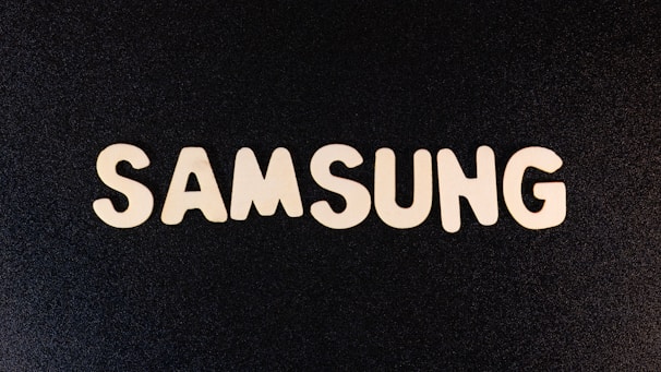The white block letters spelling 'SAMSUNG' are arranged on a textured black surface, creating a high contrast visual.