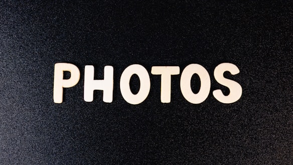 Wooden letters spell out the word 'PHOTOS' against a textured black background. The letters are evenly spaced and appear to be cut from a light-colored wood, creating a stark contrast against the dark backdrop.
