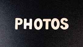 Wooden letters spell out the word 'PHOTOS' against a textured black background. The letters are evenly spaced and appear to be cut from a light-colored wood, creating a stark contrast against the dark backdrop.