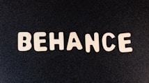 Block letters spelling 'BEHANCE' are arranged on a dark background, appearing in a casual, playful font.