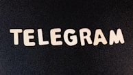 Wooden letters spelling out the word 'TELEGRAM' are arranged on a dark, textured surface.