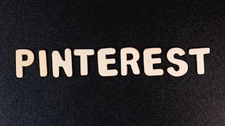 Wooden letters spell out 'PINTEREST' on a dark, textured background. The letters are evenly spaced and create a strong visual contrast against the black surface.