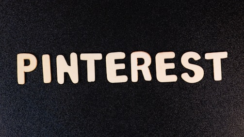 Wooden letters spell out 'PINTEREST' on a dark, textured background. The letters are evenly spaced and create a strong visual contrast against the black surface.