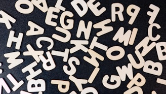 A scattered assortment of wooden alphabet letters arranged randomly on a dark surface, creating a contrast between the light wood and the black background.