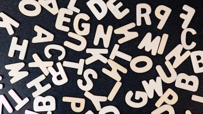 A scattered assortment of wooden alphabet letters arranged randomly on a dark surface, creating a contrast between the light wood and the black background.