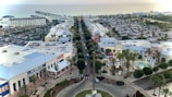 Overhead view of a coastal town featuring a main road lined with retail and entertainment buildings. There are palm trees, a large parking area filled with cars, and a pier extending into the ocean in the background. A shopping complex with bright blue and white rooftops is visible in the foreground.