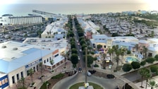 Overhead view of a coastal town featuring a main road lined with retail and entertainment buildings. There are palm trees, a large parking area filled with cars, and a pier extending into the ocean in the background. A shopping complex with bright blue and white rooftops is visible in the foreground.