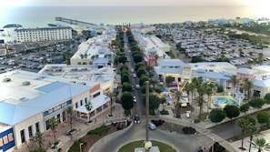 Overhead view of a coastal town featuring a main road lined with retail and entertainment buildings. There are palm trees, a large parking area filled with cars, and a pier extending into the ocean in the background. A shopping complex with bright blue and white rooftops is visible in the foreground.