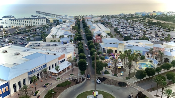 Overhead view of a coastal town featuring a main road lined with retail and entertainment buildings. There are palm trees, a large parking area filled with cars, and a pier extending into the ocean in the background. A shopping complex with bright blue and white rooftops is visible in the foreground.