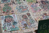 A collection of handmade artistic drawings displayed outdoors. The drawings feature various abstract and cartoonish depictions, including faces, peace symbols, hearts, and leaves. Many pieces are colorful and express messages like 'Love,' 'Create,' and 'Zen.' Some drawings incorporate elements such as cannabis leaves and a prominent image resembling a historical figure.