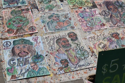 A collection of handmade artistic drawings displayed outdoors. The drawings feature various abstract and cartoonish depictions, including faces, peace symbols, hearts, and leaves. Many pieces are colorful and express messages like 'Love,' 'Create,' and 'Zen.' Some drawings incorporate elements such as cannabis leaves and a prominent image resembling a historical figure.