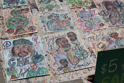 A collection of handmade artistic drawings displayed outdoors. The drawings feature various abstract and cartoonish depictions, including faces, peace symbols, hearts, and leaves. Many pieces are colorful and express messages like 'Love,' 'Create,' and 'Zen.' Some drawings incorporate elements such as cannabis leaves and a prominent image resembling a historical figure.