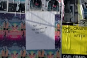 A wall covered with various street art and event posters, including a central white poster with handwritten text that reads 'THIS IS THE RECORD OF THE TIME.' The surrounding posters feature colorful designs with details of events and artists, including Carl Craig's event in black text on a bright yellow background.