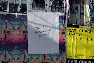 A wall covered with various street art and event posters, including a central white poster with handwritten text that reads 'THIS IS THE RECORD OF THE TIME.' The surrounding posters feature colorful designs with details of events and artists, including Carl Craig's event in black text on a bright yellow background.