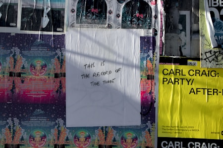 A wall covered with various street art and event posters, including a central white poster with handwritten text that reads 'THIS IS THE RECORD OF THE TIME.' The surrounding posters feature colorful designs with details of events and artists, including Carl Craig's event in black text on a bright yellow background.