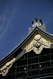 A section of a traditional Japanese roof with ornate designs, featuring intricate gold patterns and embellishments against a deep blue sky. The roof showcases detailed craftsmanship with coiled tiles and decorative gold elements.