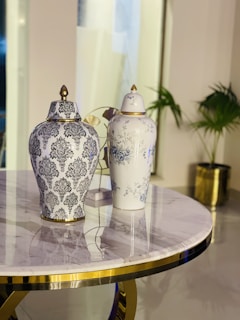 A marble-topped circular table with a gold trim is adorned with two ornate ceramic vases. One vase displays intricate black floral patterns, while the other features delicate blue floral designs. In the background, there is a potted plant with green leaves, adding a touch of nature to the decor.