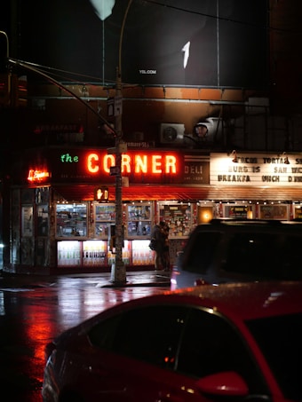 A nighttime urban scene featuring a brightly lit corner deli with a neon 'CORNER' sign. The storefront is filled with illuminated displays of drinks and snacks. A few people stand near the entrance under the glow of the lights. In the foreground, a red car is partially visible, reflecting the lights from the street. Above the deli is a large billboard showing part of a white figure or logo.