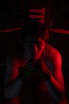 A person sits in a boxing ring, illuminated by dramatic red lighting. Their hands are wrapped with boxing tapes, and they appear deep in thought or focused. The surroundings are dark, enhancing the contrast between the shadows and the red light.