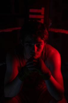 A person sits in a boxing ring, illuminated by dramatic red lighting. Their hands are wrapped with boxing tapes, and they appear deep in thought or focused. The surroundings are dark, enhancing the contrast between the shadows and the red light.