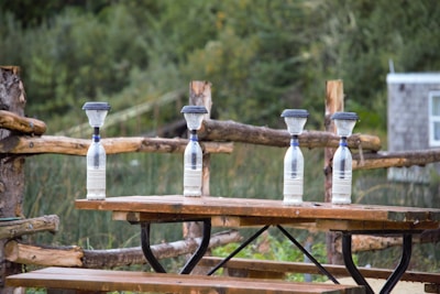 Four plastic bottles are placed on a wooden picnic table, each topped with a funnel filled with a fine material, likely sand. The table is situated outdoors with a wooden fence and lush greenery in the background, giving a serene, rustic feel.