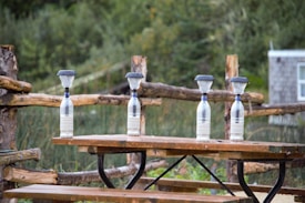 Four plastic bottles are placed on a wooden picnic table, each topped with a funnel filled with a fine material, likely sand. The table is situated outdoors with a wooden fence and lush greenery in the background, giving a serene, rustic feel.
