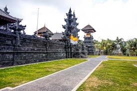 An ornate stone temple with intricate carvings stands against a cloudy sky. The structure is surrounded by a well-maintained lawn and palm trees. A pathway leads to the temple entrance, where some steps and an orange and white cloth can be seen draped across the stone pillars.