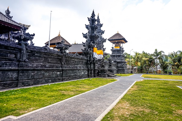 An ornate stone temple with intricate carvings stands against a cloudy sky. The structure is surrounded by a well-maintained lawn and palm trees. A pathway leads to the temple entrance, where some steps and an orange and white cloth can be seen draped across the stone pillars.