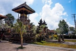 An outdoor scene featuring traditional Balinese architecture with ornate stone carvings on a building's structure and detailed rooftop. Surrounding the structure are lush green plants and trees, with bright sunlight casting shadows on the paved ground. The sky overhead is partly cloudy with patches of blue.