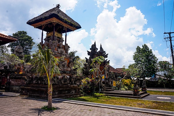 An outdoor scene featuring traditional Balinese architecture with ornate stone carvings on a building's structure and detailed rooftop. Surrounding the structure are lush green plants and trees, with bright sunlight casting shadows on the paved ground. The sky overhead is partly cloudy with patches of blue.