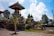 An outdoor scene featuring traditional Balinese architecture with ornate stone carvings on a building's structure and detailed rooftop. Surrounding the structure are lush green plants and trees, with bright sunlight casting shadows on the paved ground. The sky overhead is partly cloudy with patches of blue.
