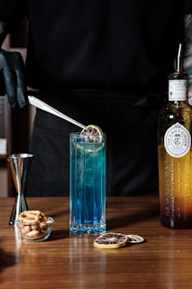 A person wearing black gloves uses tweezers to place a dried citrus slice into a tall, blue cocktail glass. Beside the glass is a bottle labeled as ginger liqueur with a metal straw inside it. A small glass bowl filled with mixed nuts and a jigger are also placed on the wooden surface. There are additional dried citrus slices on the table.