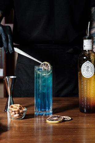 A person wearing black gloves uses tweezers to place a dried citrus slice into a tall, blue cocktail glass. Beside the glass is a bottle labeled as ginger liqueur with a metal straw inside it. A small glass bowl filled with mixed nuts and a jigger are also placed on the wooden surface. There are additional dried citrus slices on the table.