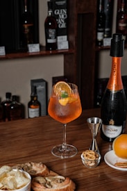 A wooden table is set with an assortment of items including a glass of Aperol Spritz garnished with an orange slice, a bottle of Cinzano Prosecco, a jigger, an orange, dried orange slices and some small snacks such as pretzels and nuts. In the background, there are several bottles of liquor displayed on shelves.