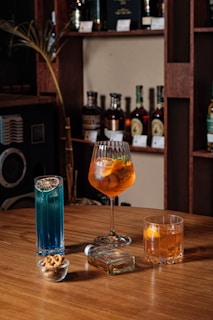 Three different types of cocktails in distinct glasses are placed on a wooden tabletop. Behind them, bottles of alcohol are arranged neatly on wooden shelves against the wall. There is a blue cocktail with a slice of dried lime, an orange cocktail in a large goblet glass with slices of orange and other fruits, and a shorter glass with another orange drink containing citrus slices. Additionally, a small glass bowl with a few pretzels and a clear glass bottle also sit on the table.